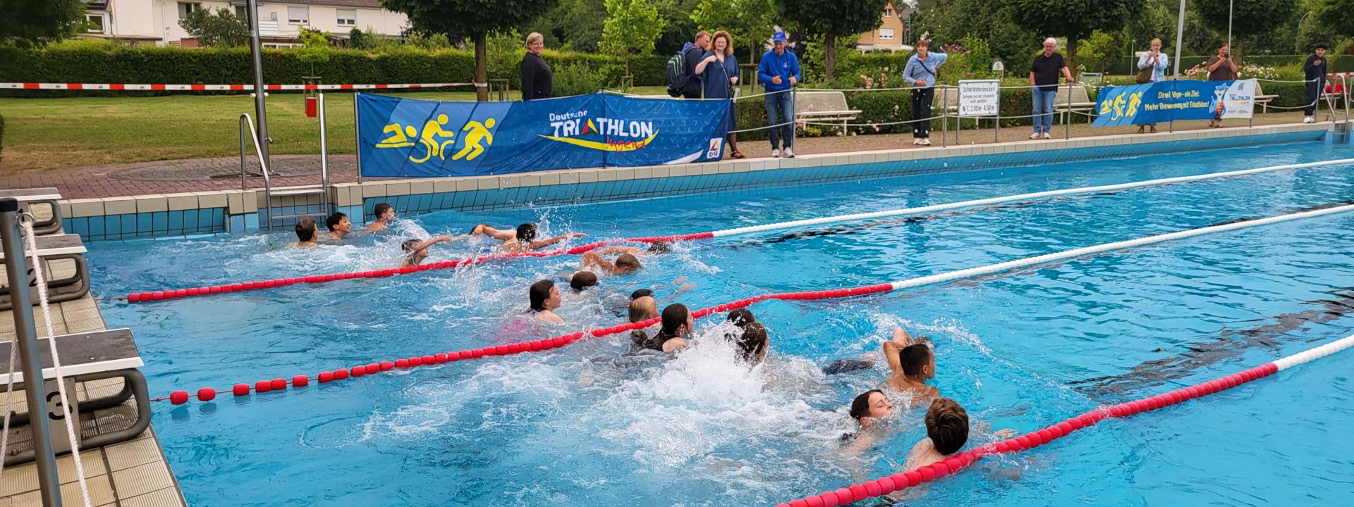 Kinder und Jugendliche schwimmen auf Bahnen in einem Freibad während einer Triathlon-Veranstaltung, beobachtet von Zuschauern am Beckenrand.