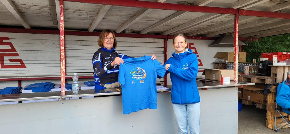 Zwei lächelnde Frauen stehen an einem Stand und halten gemeinsam ein blaues Event-T-Shirt mit der Aufschrift „Volksbank Schultriathlon Ahlen“ in die Kamera.