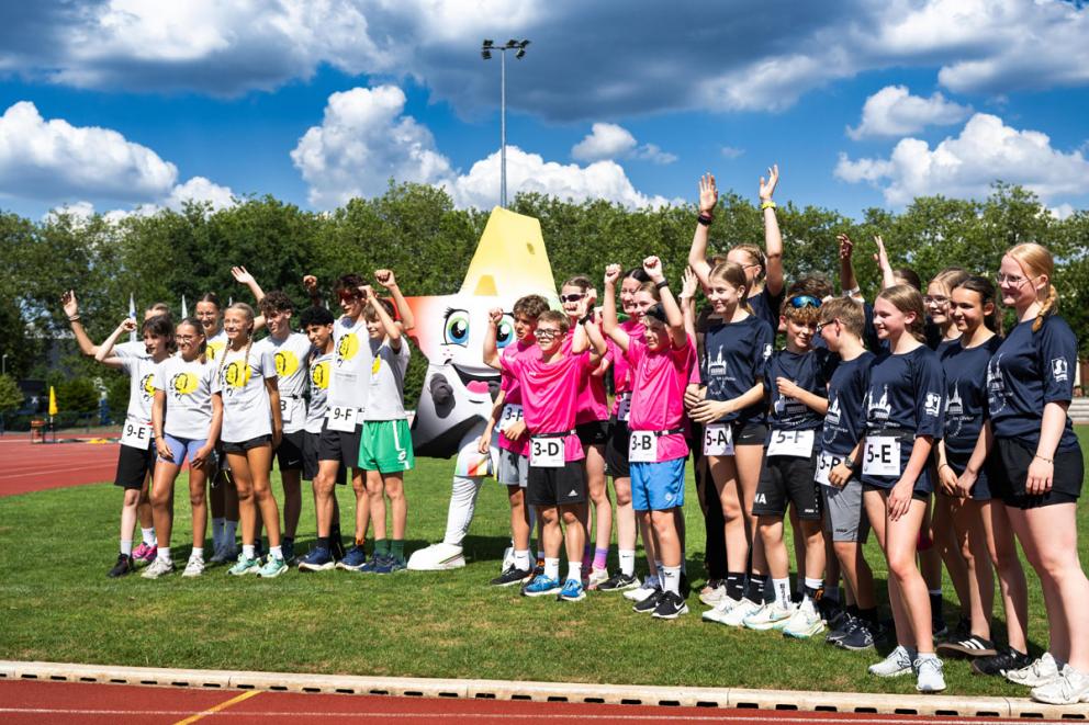 Auf dem Bild sind drei gemischte Sportmannschaften zu sehen, die in die Kamera winken. In der Mitte steht ein lebendes Maskottchen in Form eines Sternes. 