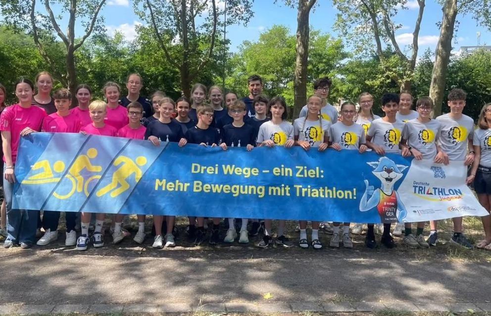 Mehrere Kinder und Jugendliche stehen hinter einem Banner mit dem Slogan: "Drei Wege, ein Ziel! Mehr Bewegung mit Triathlon!"